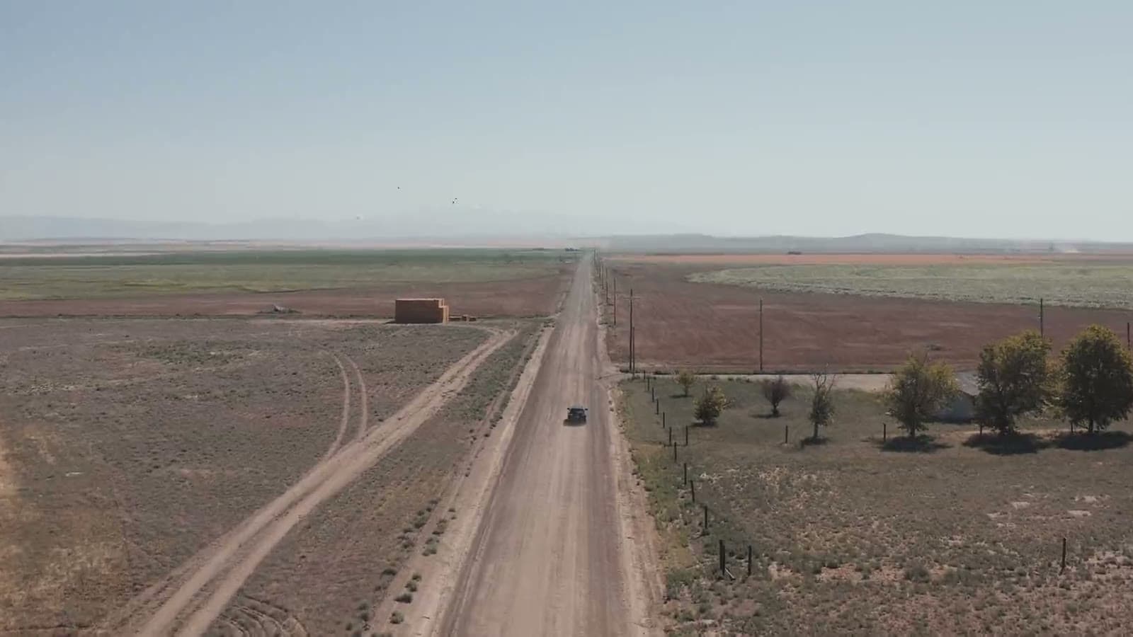 A car driving down a long dirt road through dry farmland, seen from a drone