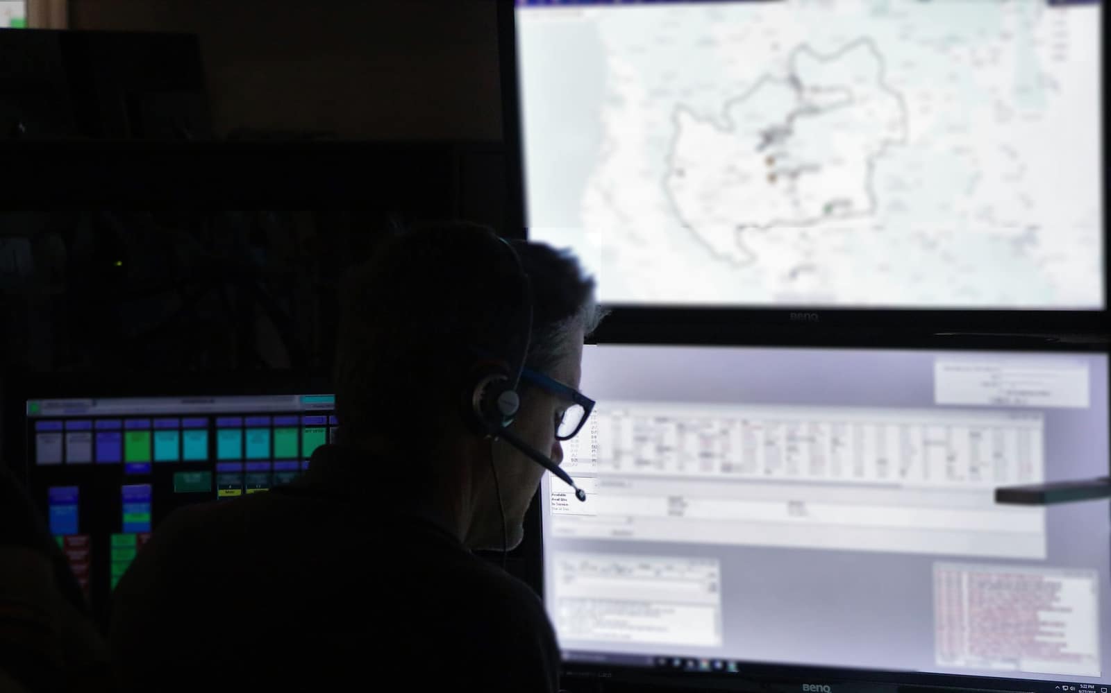 Un despachador con audífonos, visto a contraluz, observa un mapa en una pantalla grande dentro de un centro de despacho a oscuras